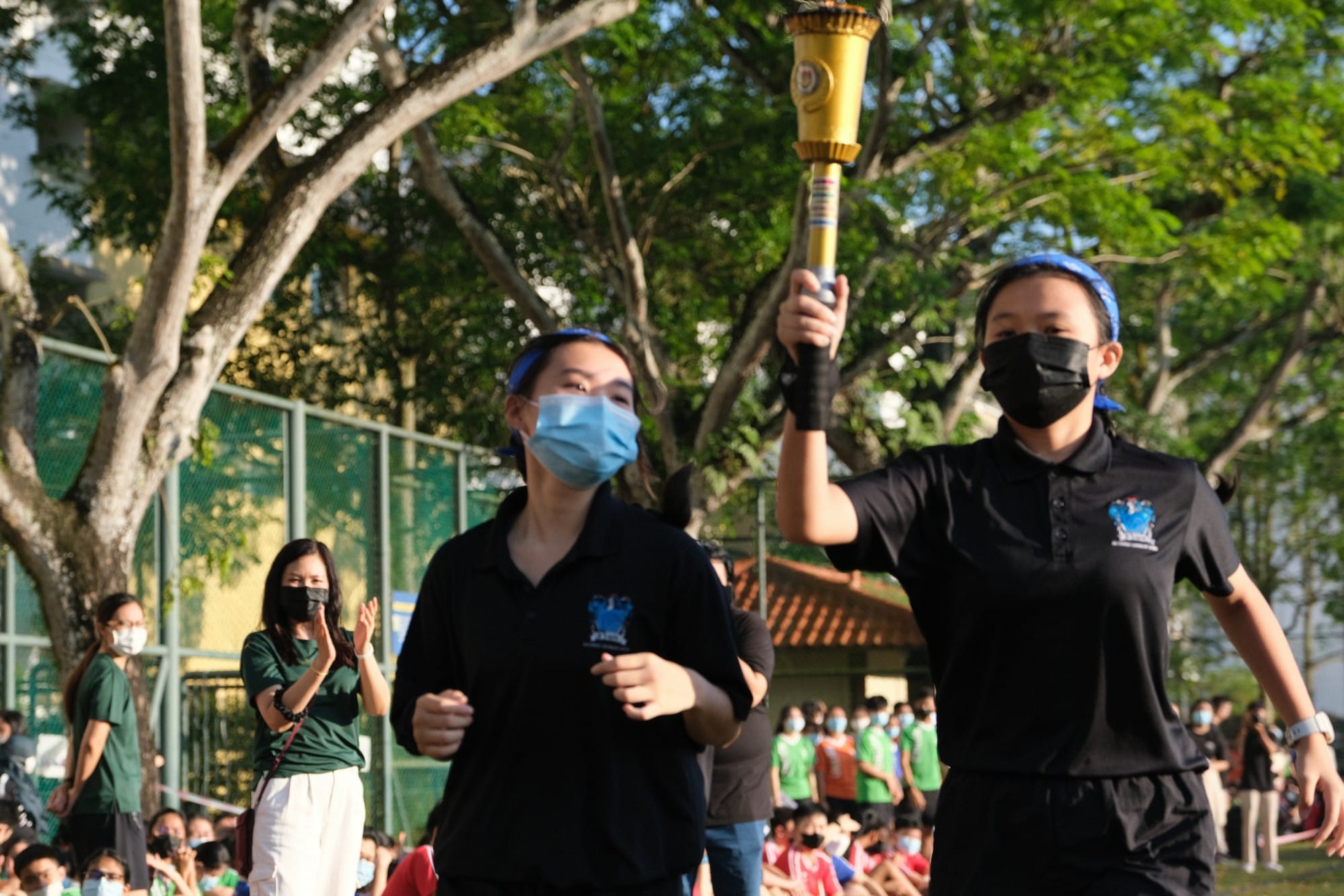 Two masked students in black uniforms carry a golden torch while running, with a crowd watching outdoors.
